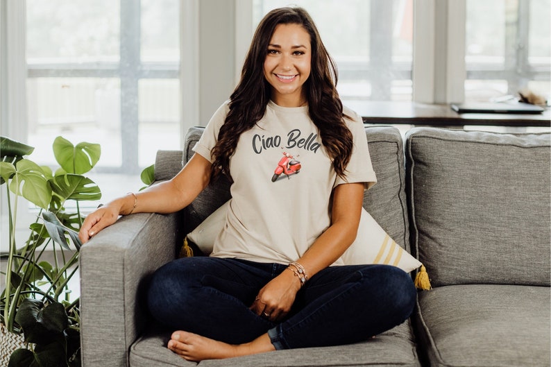 May include: A woman wearing a beige t-shirt with the text "Ciao Bella" and a red scooter graphic. She is sitting on a gray couch with a white pillow and a yellow striped pillow.