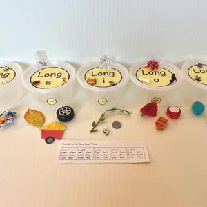 May include: Five clear plastic containers labeled Long a, Long e, Long i, Long o, and Long u, each containing small objects. A white paper card lists the contents of the Long Vowel Tubs, including items like a chain, leaf, tiger, rose, and cube.