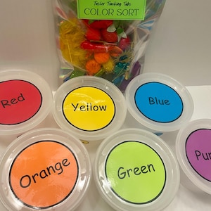 May include: Six clear plastic containers with circular labels in red, yellow, blue, orange, green, and purple, each featuring the corresponding color name in black text. Behind the containers is a clear bag labeled Tusler Teaching Tubs COLOR SORT.