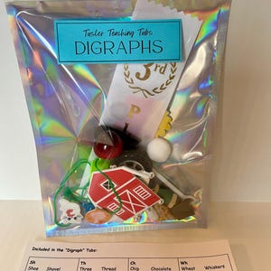 May include: A clear plastic bag filled with various small objects, including a red barn, a white cotton ball, a green ribbon, and a red and white ball. The bag is labeled "Tusler Teaching Tubs DIGRAPHS 3rd". A white card with a chart lists digraphs and words that use them, such as "sh, shoe, shovel, shell, shed, th, three, thread, thumb, thunder, third, thimble, ch, chip, chocolate, chain, cherry, chicken, wh, wheat, whiskers, whale, white, wheel."