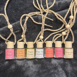Op de afbeelding: Een verzameling van zeven kleine, heldere glazen flesjes met houten doppen, elk gelabeld met een andere geurnaam. De flesjes hangen aan beige koorden met houten kralen. De labels bevatten namen als "Pink Sugar", "Ginger Bread Cookie" en "Marshmallow Fluff".
