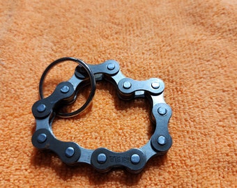 May include: A black metal keychain shaped like a heart, made of linked bicycle chain. The keychain has a small metal ring attached to the top.
