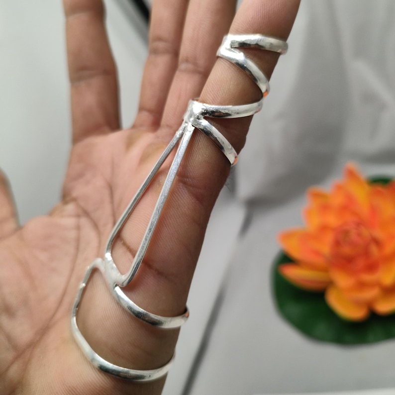 Op de afbeelding: Een zilveren vingerring met een uniek ontwerp. De ring heeft meerdere banden en verbindingsstaven, waardoor een ingewikkeld en opvallend accessoire ontstaat. De ring wordt om een vinger gedragen en toont zijn gedetailleerde vakmanschap en moderne esthetiek.