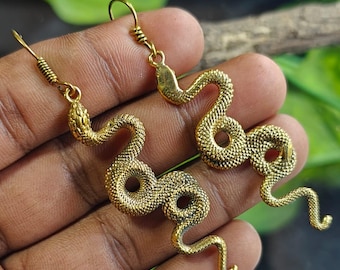 May include: A pair of gold-colored snake earrings. The earrings are detailed with scales and have a curved, serpentine design. The earrings are shown against a blurred green background.