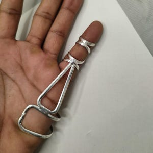 Op de afbeelding: Een zilveren vingerring met een uniek ontwerp. De ring heeft drie verbonden banden die rond de vinger gaan, met een geometrisch ontwerp dat zich langs de vinger uitstrekt. De ring is gemaakt van een glanzend, metalen materiaal.