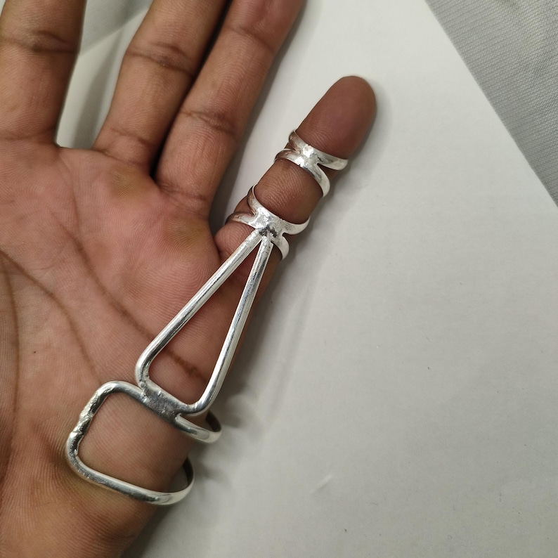 Op de afbeelding: Een zilveren vingerring met een uniek ontwerp. De ring heeft drie verbonden banden die rond de vinger gaan, met een geometrisch ontwerp dat zich langs de vinger uitstrekt. De ring is gemaakt van een glanzend, metalen materiaal.