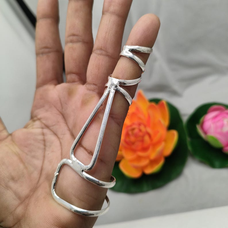 Op de afbeelding: Een zilveren vingerpantserring met een uniek ontwerp. De ring heeft meerdere banden en een decoratief element dat zich over de vinger uitstrekt. De ring wordt getoond op een hand met een vage achtergrond van bloemen.