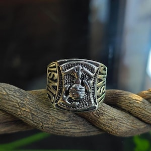 May include: A silver ring with the letters "USMC" on the sides. The ring features a detailed emblem with an eagle, anchor, and globe design. The ring is displayed on a brown rope.