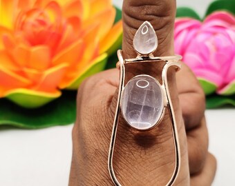 May include: A rose gold finger ring with three rose quartz stones. The ring features a teardrop-shaped stone, a large oval stone, and a smaller ring at the top of the finger. The ring is displayed against a background of orange and pink flowers.