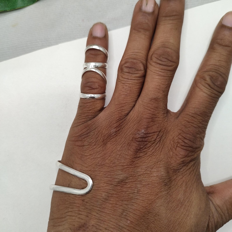 Op de afbeelding: Een hand met meerdere zilveren ringen en een zilveren manchetarmband. De ringen hebben verschillende ontwerpen, waaronder een eenvoudige band en een ring met een verbindend ontwerp. De manchetarmband heeft een U-vormig ontwerp.