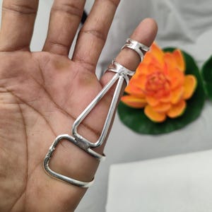 Op de afbeelding: Een zilveren vingerringset. De set bevat een ring voor de basis van de vinger, een ring voor het midden en een ring voor de vingertop, allemaal verbonden door een zilveren frame. De achtergrond bevat een oranje bloem en een groen blad.
