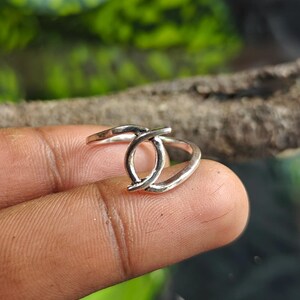 May include: A silver ring with a unique design. The ring features two curved bands that intersect, creating an open, abstract shape. The ring is shown on a finger, with a blurred green background.