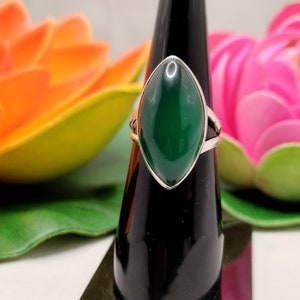 May include: A silver ring featuring a large, oval-shaped, deep green gemstone. The gemstone is set in a silver band with a split shank design. The ring is displayed on a black stand, with colorful flowers in the background.