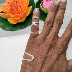 Op de afbeelding: Een hand met zilveren ringen en een manchetarmband. De ringen omvatten een eenvoudige band, een ring met dubbele band en een ring met een X-vormig ontwerp. De manchetarmband heeft een U-vormig ontwerp.