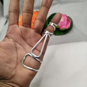 Op de afbeelding: Een zilveren vingerpantserring met een geometrisch ontwerp. De ring heeft een driehoekige vorm en meerdere banden die om de vinger wikkelen. De ring wordt gedragen op een hand met een vage achtergrond.