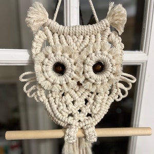 May include: A macrame owl wall hanging in cream-colored cotton rope. The owl features large, round eyes with brown wooden beads, a wooden perch, and decorative tassels. The owl is approximately 12 inches tall.