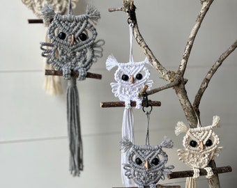 Puede incluir: Cinco adornos de búho de macramé colgando de ramas. Los búhos están hechos con hilo blanco, gris y beige. Cada búho tiene una percha de madera y un borla que cuelga de la parte inferior.