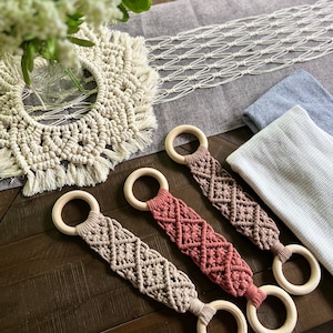 May include: Three wooden ring teethers with macrame knotting in beige and terracotta colors. The teethers are on a wooden surface.