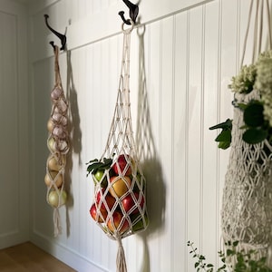 May include: Three macrame hanging baskets with a white background. The baskets are filled with apples, onions, and flowers. The baskets are hanging from hooks on a white wall.