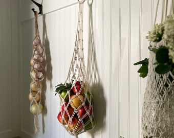 May include: Three macrame hanging baskets with a white background. The baskets are filled with apples, onions, and flowers. The baskets are hanging from hooks on a white wall.