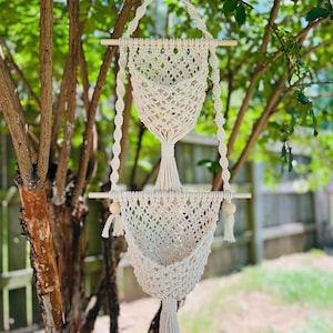 May include: A white macrame plant hanger with two hanging baskets and wooden dowels. The hanger is made of cotton cord and has a tassel at the bottom.