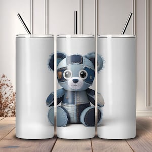 May include: White insulated tumbler with a black straw, featuring a patchwork denim teddy bear design. The bear is in shades of blue and has a stitched appearance. The tumbler is designed for cold beverages.