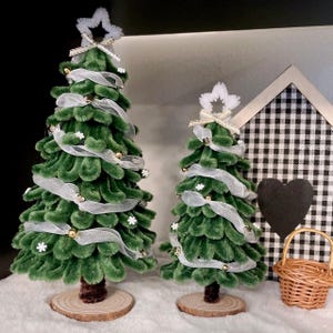May include: Two green velvet pipe cleaner Christmas trees decorated with white ribbon, gold beads, and white snowflakes. The trees are on wooden bases and are sitting on a white surface.