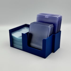 May include: A blue card holder with three compartments. Two compartments hold stacks of clear card sleeves, and the third holds a stack of blue cards. The text "PJE" is visible on the card sleeves.