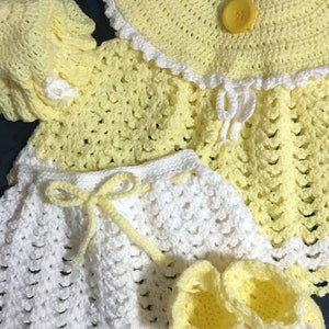 May include: A handmade crochet baby outfit in yellow and white. The set includes a dress, pants, bonnet, and booties. The dress has a yellow collar with a white trim and a yellow button. The pants and booties are white with yellow accents.