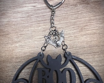Pode incluir: Chaveiro preto com tema de Halloween com um design de morcego e a palavra "BOO". O chaveiro tem um porta-chaves prateado em forma de coração e uma corrente com um pingente de morcego. As asas do morcego estão abertas.