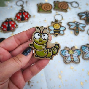 May include: A ceramic caterpillar figurine with a glossy green glaze, large white eyes, and black outlines. The piece features a segmented body and two antennae. Other insect-themed ceramic charms, including a ladybug, snail, bee, and butterfly, are visible in the background.