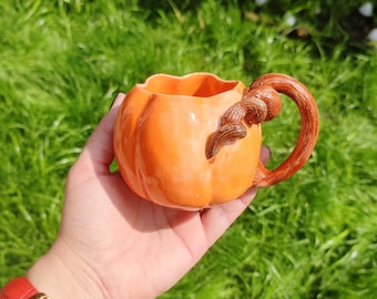 May include: Orange ceramic pumpkin mug with a brown handle shaped like a stem. The mug is sitting on a hand.