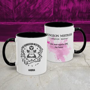 May include: Two white ceramic mugs with black handles and rims. One mug features a black and white graphic with the text "THE DUNGEON MISTRESS" and "AABRIA". The other mug has the text "DUNGEON MISTRESS" and "Like your regular DM, but better."