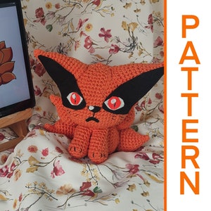 May include: A crocheted orange and black fox plushie with red eyes. The plushie is sitting on a floral patterned fabric. The image also shows a tablet with a cartoon image of the fox on the screen. The text "PATTERN" is written vertically in orange on the right side of the image. The text "Italiano" is written vertically in orange on the right side of the image.