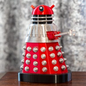 Puede incluir: Una figura de Dalek roja y plateada con una base negra. El Dalek tiene una cabeza en forma de cúpula con un ojo negro y un cuerpo plateado con un arma plateada.