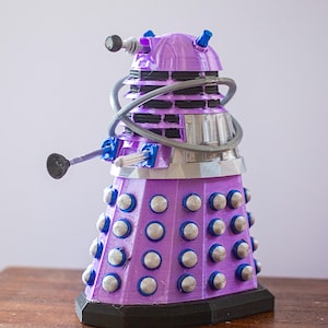 Könnte beinhalten: Eine lila Dalek-Figur mit silbernen, schwarzen und blauen Akzenten. Die Science-Fiction-Figur hat einen zylindrischen Körper mit silbernen Kugeln und einen gewölbten Kopf. Sie verfügt über einen pistolenartigen Arm und einen Manipulatorarm und steht auf einem schwarzen Sockel.