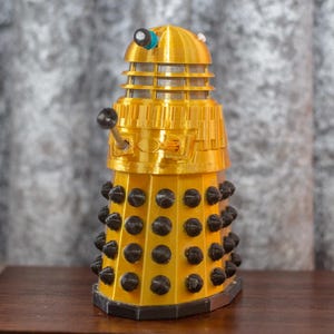 Op de afbeelding: Een gouden Dalek-figuur met zwarte details. De Dalek heeft een koepelvormige kop met een rasterpatroon en een zwart oog. Het lichaam is bedekt met zwarte cirkels.