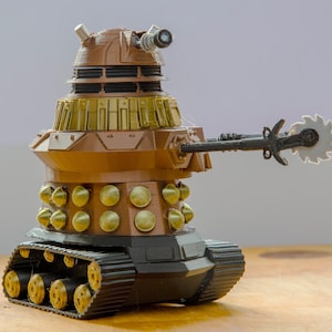 Könnte beinhalten: Ein braunes und goldenes Spielzeug-Dalek mit einer sägeartigen Waffe. Das Spielzeug hat eine Panzerbasis mit schwarzen Ketten und goldenen Rollen. Der Körper des Dalek weist goldene Akzente und einen kanonenartigen Anhang auf.