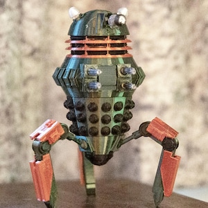 May include: A detailed model of a Dalek, featuring a green and orange colour scheme. The model has a cylindrical body with black spheres and orange accents. It stands on three legs and has two white lights on top.