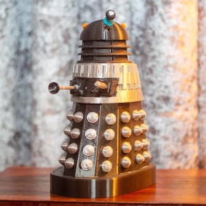 Könnte beinhalten: Eine schwarze und silberne Dalek-Figur mit einem kuppelförmigen Kopf, einem silbernen Kragen und einem schwarzen Sockel. Der Dalek hat ein einzelnes Auge und eine kleine, schwarze Waffe an der Seite.