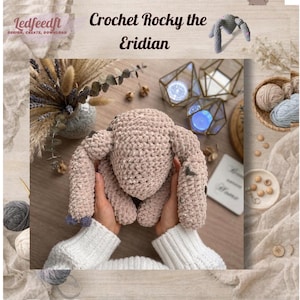 Peut inclure: Un jouet en forme de lapin en crochet rose clair, avec de longues oreilles et une texture douce. L'image comprend le texte "Crochet Rocky the Eridian" et le logo "ledfeedft DESIGN, CREATE, DOWNLOAD". Le lapin est tenu dans les mains.