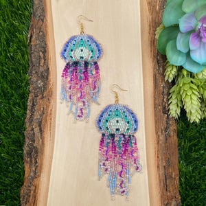 May include: A pair of beaded jellyfish earrings. The earrings feature a blue, teal, and purple beaded top with dangling strands of pink, purple, and blue beads. The earrings are displayed on a wooden surface.