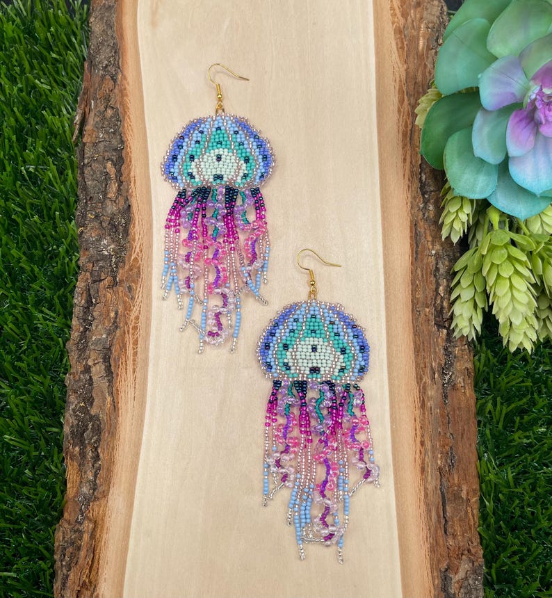 May include: A pair of beaded jellyfish earrings. The earrings feature a blue, teal, and purple beaded top with dangling strands of pink, purple, and blue beads. The earrings are displayed on a wooden surface.