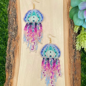 May include: A pair of beaded jellyfish earrings. The earrings feature a blue, teal, and white beaded top with dangling strands of pink, purple, and blue beads. The earrings have gold-colored ear wires and are displayed on a wooden surface.