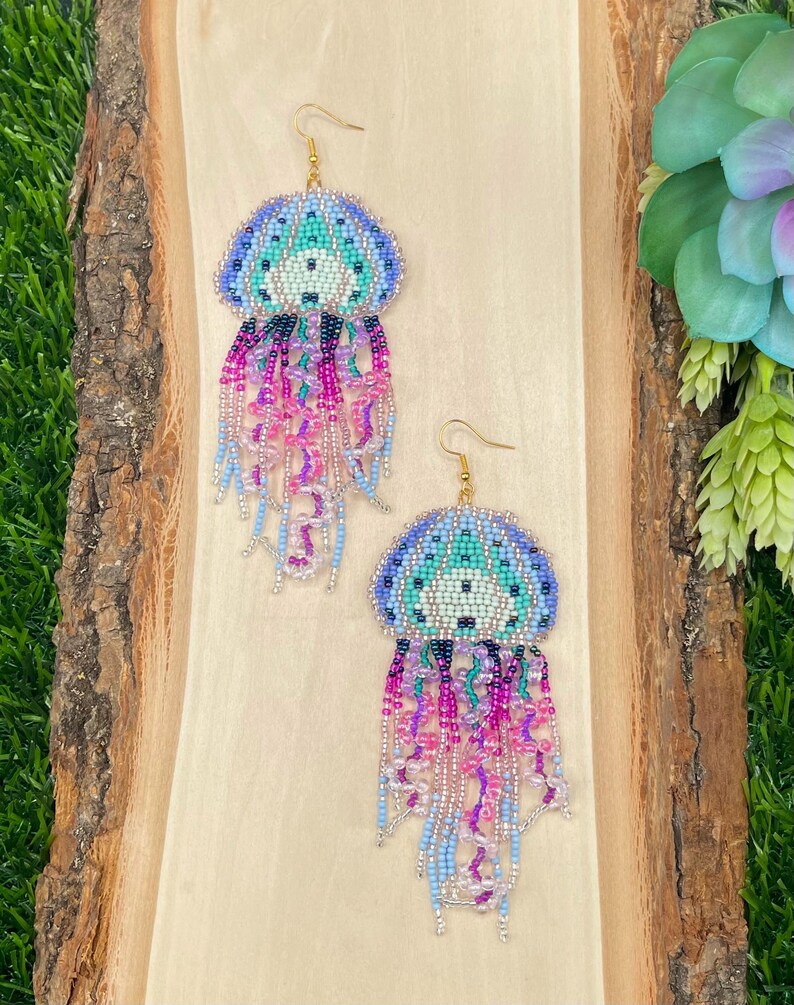 May include: A pair of beaded jellyfish earrings. The earrings feature a blue, teal, and white beaded top with dangling strands of pink, purple, and blue beads. The earrings have gold-colored ear wires and are displayed on a wooden surface.