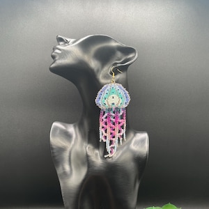 May include: Handmade beaded jellyfish earrings. The earrings feature a detailed design with a blue, teal, and white top, and pink, purple, and silver dangling strands. The earrings are displayed on a black mannequin head.