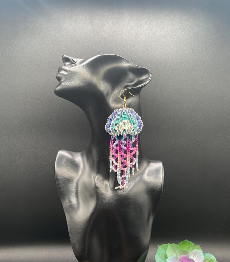 May include: Handmade beaded jellyfish earrings. The earrings feature a detailed design with a blue, teal, and white top, and pink, purple, and silver dangling strands. The earrings are displayed on a black mannequin head.