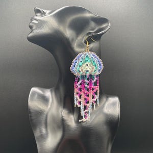 May include: Handmade beaded jellyfish earrings. The earrings feature a dome-shaped top in shades of blue, teal, and white, with a central design. Long, dangling strands in pink, purple, and blue create the jellyfish tentacles. Gold-colored ear hooks.