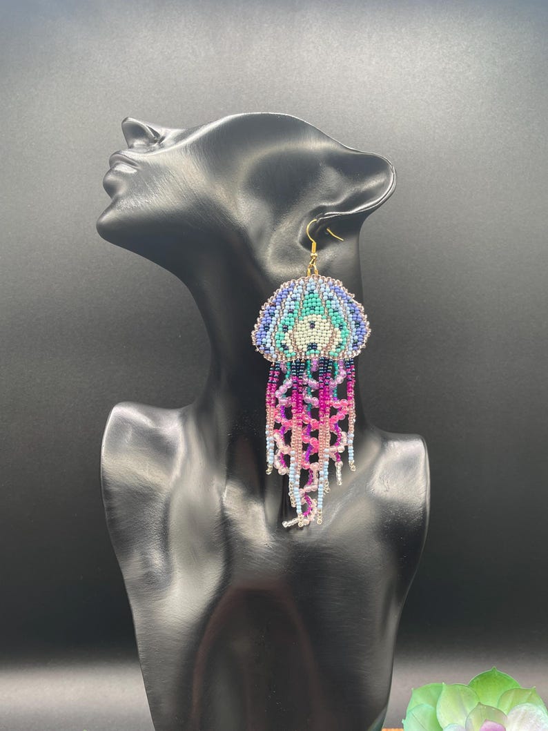 May include: Handmade beaded jellyfish earrings. The earrings feature a dome-shaped top in shades of blue, teal, and white, with a central design. Long, dangling strands in pink, purple, and blue create the jellyfish tentacles. Gold-colored ear hooks.