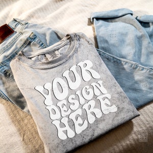 Puede incluir: Una camiseta gris con un gráfico blanco que dice "YOUR DESIGN HERE" en una fuente retro. La camiseta está doblada y colocada encima de un par de jeans azul claro.